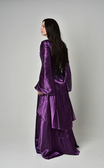 full length portrait of beautiful girl with long black hair,   wearing purple fantasy medieval gown. standing pose with back to the camera on grey studio background.