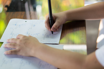 Close up kid's hand writing on paper, writing messy math on wooden table in room,student child girl holding pen doing homework at home, calculate the results on paper , education concept