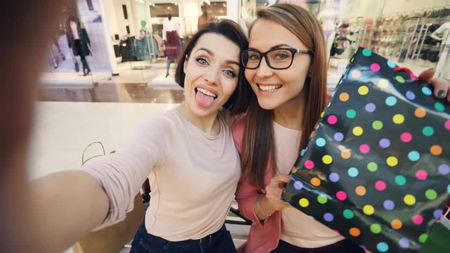 Point of view shot of pretty girls friends taking selfie with paper bags in shopping mall and having fun showing tongue and making funny faces. Friendship and technology concept.