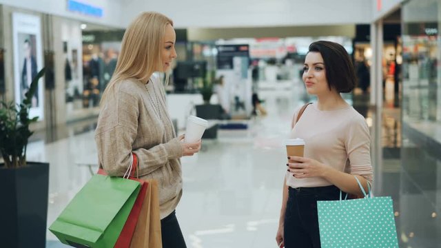 Beautiful young women are chatting in shopping mall holding takeaway drinks and paper bags discussing purchases and sharing news. Youth lifestyle and stores concept.