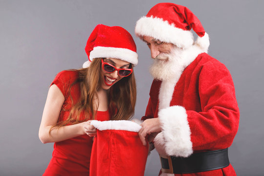 Santa Claus with white beard wearing sungasses and young mrs. Claus wearing Santa hat, red dress and sunglasses standing on the gray background, surprised mrs. Santa searching some presents in the