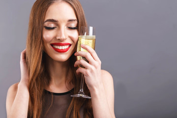Fototapeta premium Portrait of awesome long haired woman with red lips wearing black dress holding glass of champagne near her face, New Year, Christmas, holidays
