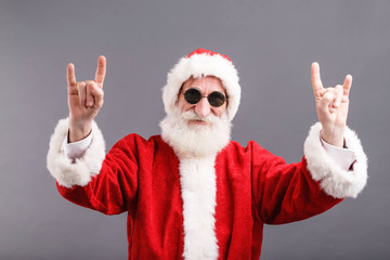 Portrait of Santa Claus with a white beard wearing sunglasses and Santa outfit standing and showing a rock gesture on the gray background, New Year, Christmas, holidays, souvenirs, gifts, shopping