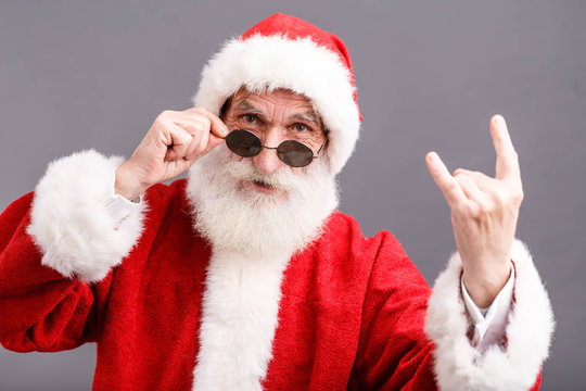 Portrait of Santa Claus with a white beard wearing sunglasses and Santa outfit standing and showing a rock gesture on the gray background, New Year, Christmas, holidays, souvenirs, gifts, shopping