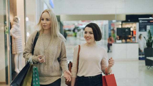 Attractive blonde is shopping with her pretty female friend chatting and smiling walking in mall then looking at underwear in shop window and talking.
