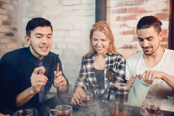 Happy friends relaxing in loft bar smoking hookah. Young man pours tea from thermos