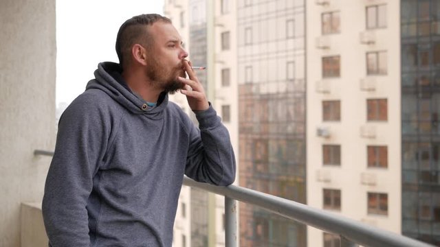 The guy with the beard is standing on the balcony smoking a cigarette