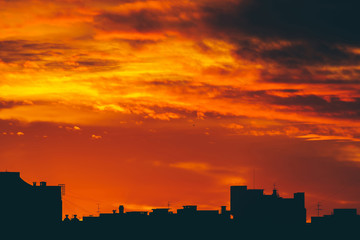 Cityscape with vivid fiery dawn. Amazing warm dramatic cloudy sky above dark silhouettes of city buildings. Orange sunlight. Atmospheric background of sunrise in overcast weather. Copy space.