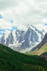 Obraz premium Snowy mountain top behind rocky mountains under cloudy sky. Rocky slope above forest. Clouds above glacier. Atmospheric landscape of majestic nature of highlands.