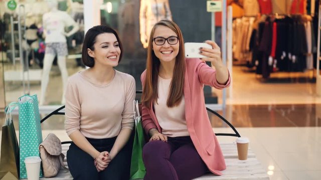 Cute girls are making online video call using smartphone sitting on bench in shopping mall talking, looking at phone camera and showing purchases in paper bags.