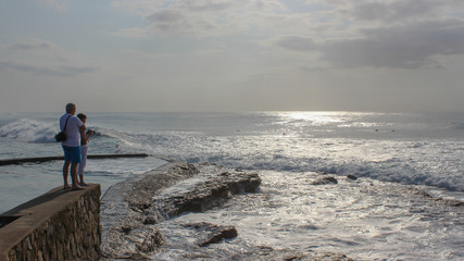 middle aged couple standing on a pier watching the waves crashing over rocks at sunrise 