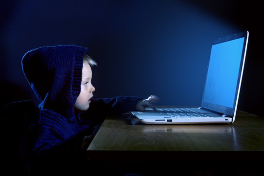A little boy in a black hood uses a laptop in the dark.