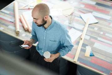 Directly above view of serious handsome young bearded designer in blue shirt standing at desk and checking message on smartphone while drinking coffee at workplace