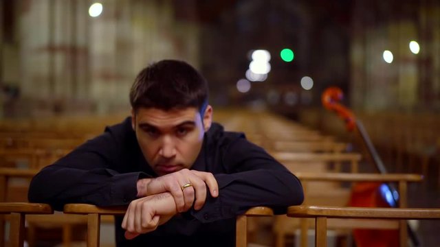 sad man is sitting bowing head on crossed hands on chair in empty hall of church, thinking