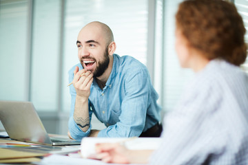 Cheerful excited handsome male designer leaning on table with open laptop and color swatches and laughing during meeting with colleague