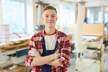 Content confident handsome young carpentry student in work gloves wearing red checkered shirt and overall standing in modern workshop and looking at camera