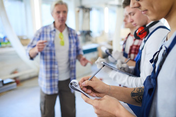 Close-up of young interns standing in row and making notes while listening to carpenter, content guy with tattoo writing out information in clipboard