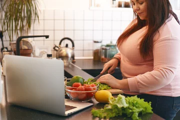 Poster Übergewichtige Frau, die einen Laptop verwendet, um ein Videorezept anzuschauen, während sie veganen Vitamin-Avocado-Salat zubereitet und Blattsalat auf einem Holzbrett schneidet. Gesundes Essen, Gewichtsverlust, Diät und Ernährungskonzept © shurkin_son