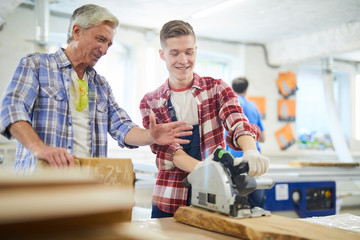 Positive young carpentry student in work gloves standing at workbench and using circular saw under...