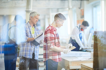 Serious concentrated mature foreman in casual shirt holding clipboard making notes while controlling young worker who making design on wooden plank