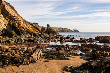 Irish landscape with a rocky beach and a distant lighthouse on a summer day. Baily Lighthouse as seen from the Howth Cliff Walk, Dublin, Ireland.