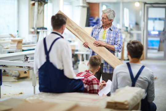 Confident handsome mature male carpenter in casual shirt standing in front of student group and holding wooden plank while teaching students to process wood