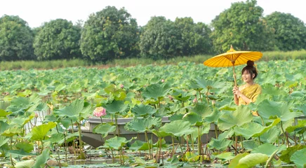Fotobehang Lotusbloem Beautiful asia women wearing traditional Thai dress and sitting on wooden boat with yellow paper umbrella in flower lotus lake. Her hands are holding a umbrella . And her face is be happy and smile.  © Prot