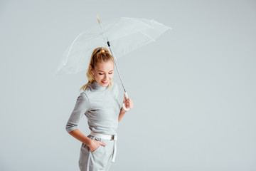 smiling woman in grey clothes and hand in pocket posing with transparent umbrella isolated on grey