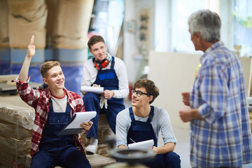 Content handsome blonde guy in overall sitting on stack of packaged planks and raising hand while having question at carpentry class - obrazy, fototapety, plakaty