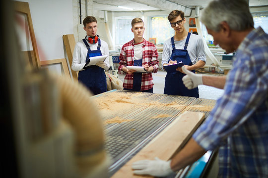 Professional senior carpenter in gloves showing thumb-up while explaining young students how to work with woods, he checking surface of plank placed on rubber mat in workshop