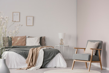 Wooden armchair next to bed with blanket and cushions in white bedroom interior. Real photo