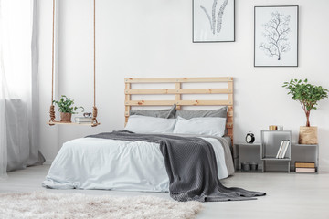 Stylish swing with books and plant in pot next to king size bed with wooden headboard and grey and white bedding - obrazy, fototapety, plakaty