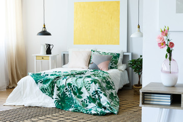 Abstract golden painting on the white wall of trendy bedroom interior with floral bedding on cozy bed and jugs on nightstand