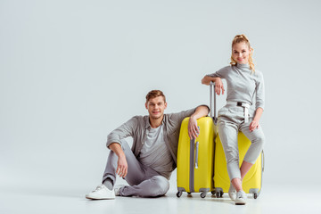 couple sitting near yellow suitcases and looking at camera on grey background, travel concept