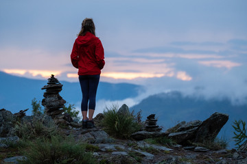 Obraz premium Young woman in red stands near the cairns and looks into the valley at sunset. Altai Republic, Russia. Image has some noise
