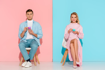Obraz premium couple sitting on chairs, holding toy coffee cups and looking at camera on pink and blue background