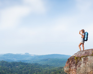 Young woman hiker stands with backpack on the cliff and looks into valley