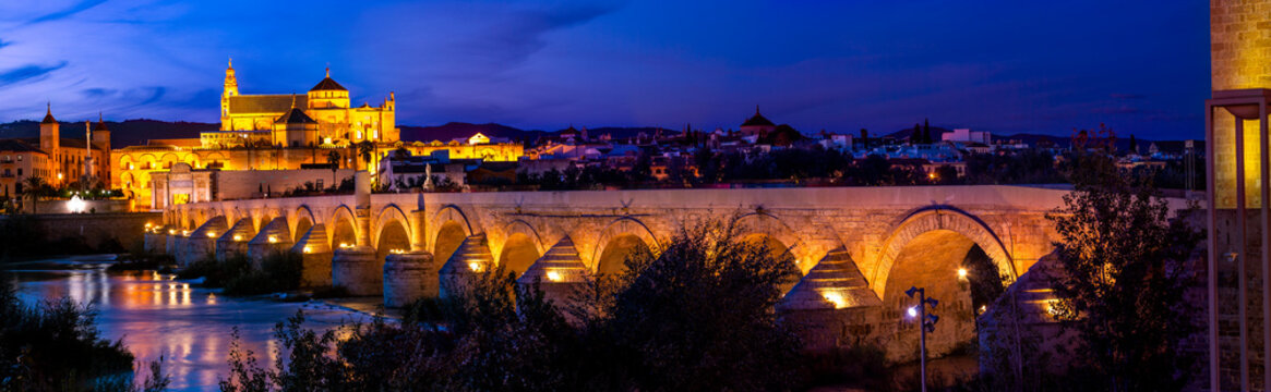 Panorama of ancient stone Roman bridge illuminated in night time with glowing Moorish Mosque Cathedral (Mezquita) on background in Cordoba, Spain