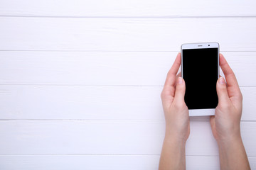 Female hands holding mobile phone with blank screen on white wood background. Smartphone on wood table.