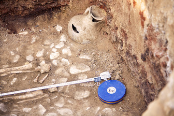 Real archaeological excavations. The remains of human bones in the tomb and well-preserved clay jar found in the ground. Outdoors, close up, copy space.