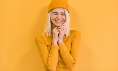 Cheerful blonde female with positive expression, close her eyes and smiling joyfully, wears yellow clothes, isolated over yellow background. Happy woman feeling excited. People and emotions concept.