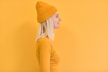 Profile portrait of happy blonde hair woman wears yellow clothes, hat, smiles positively, isolated over yellow background. Copy space for advertisement or promotion. Caucasian female stands sideways