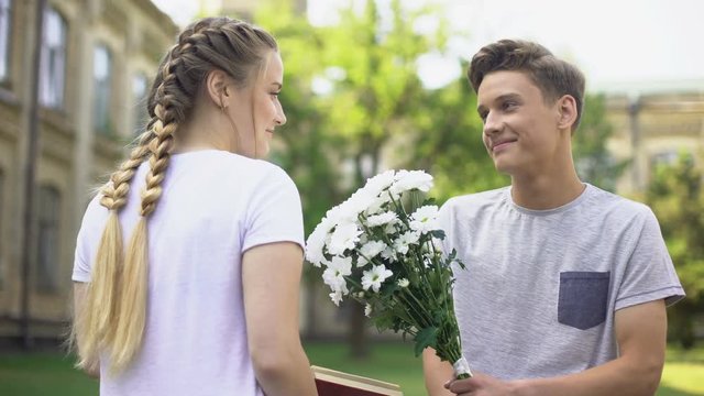 Teen boy presenting field flowers to teen girl, reading book in city garden