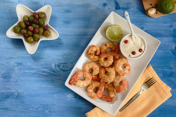 Delicious fried shrimp on a white plate next to lime and garlic sauce on a blue wooden background next to olives and a napkin