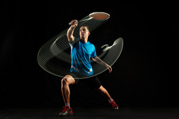 Young man playing badminton over black studio background. Fit male athlete isolated on dark with led light trail . badminton player in action, motion, movement. attack and defense concept - obrazy, fototapety, plakaty