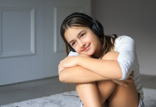 Beautiful happy romantic smiling preteen girl with wireless headphones listening to music sitting on floor at home hugging her khees relaxing. Calm leisure time