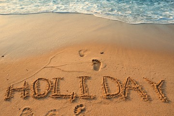 Concept HOLIDAY handwriting on sand beach with waves and blue ocean.