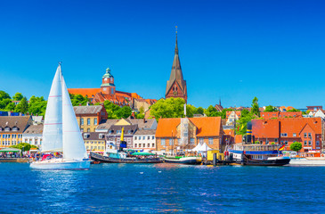 Cityscape of Flensburg. Panorama of a small European town in Northern Germany. A sailboat is floating in a harbour along the coastline with old architecture, ships and landmarks on the background