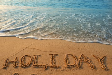 Concept HOLIDAY handwriting on sand beach with waves and blue ocean.
