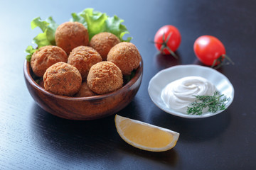 Falafel lies in a wooden plate. On the table lie tomatoes, cucumbers, lettuce, dill, lemon, sour cream.